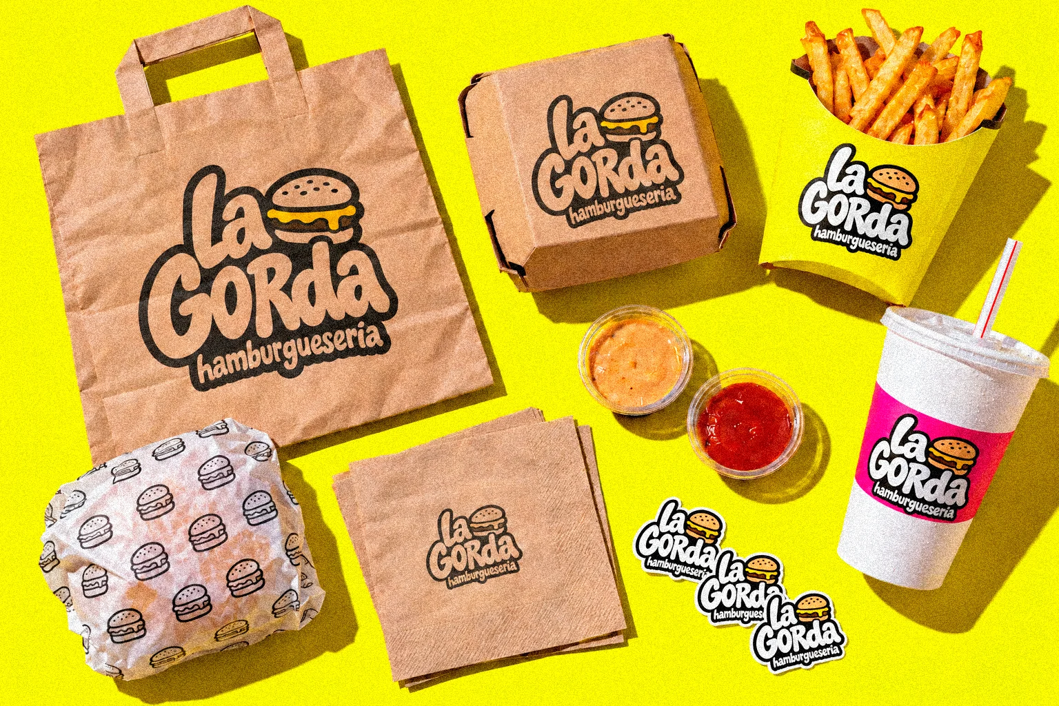 Bolsa, caja, patatas, vaso, servilletas y pegatinas de La Gorda vistos desde arriba sobre fondo amarillo