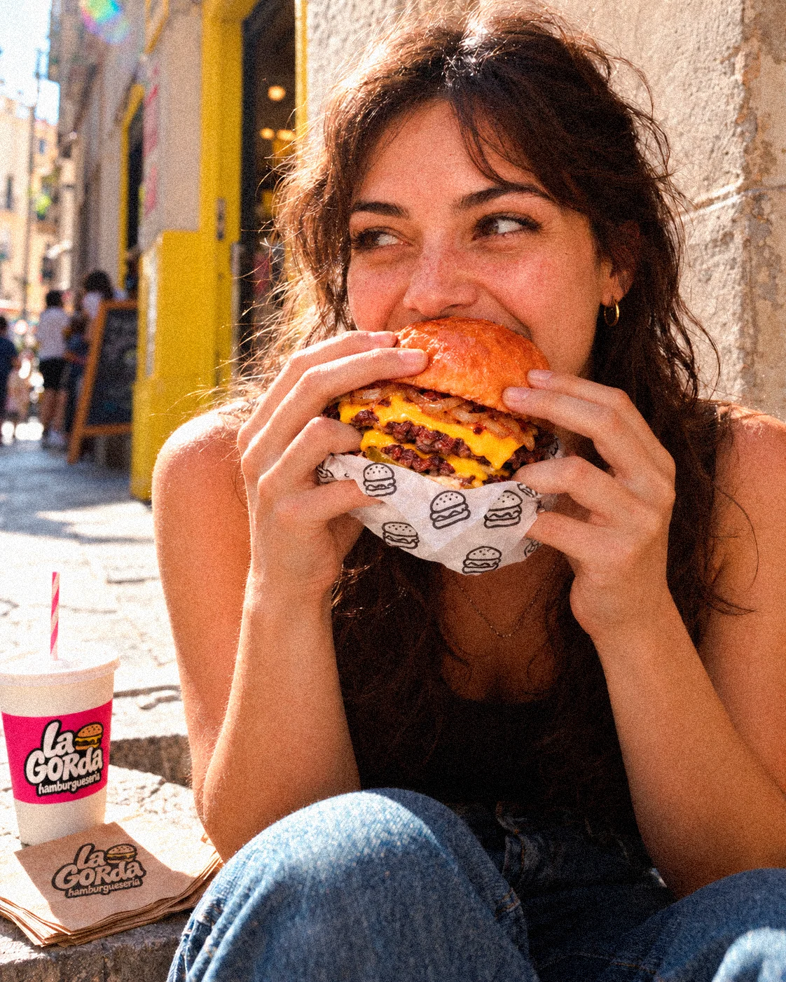Una chica come una hamburguesa La Gorda sentada en una calle soleada, con el vaso de la marca al lado