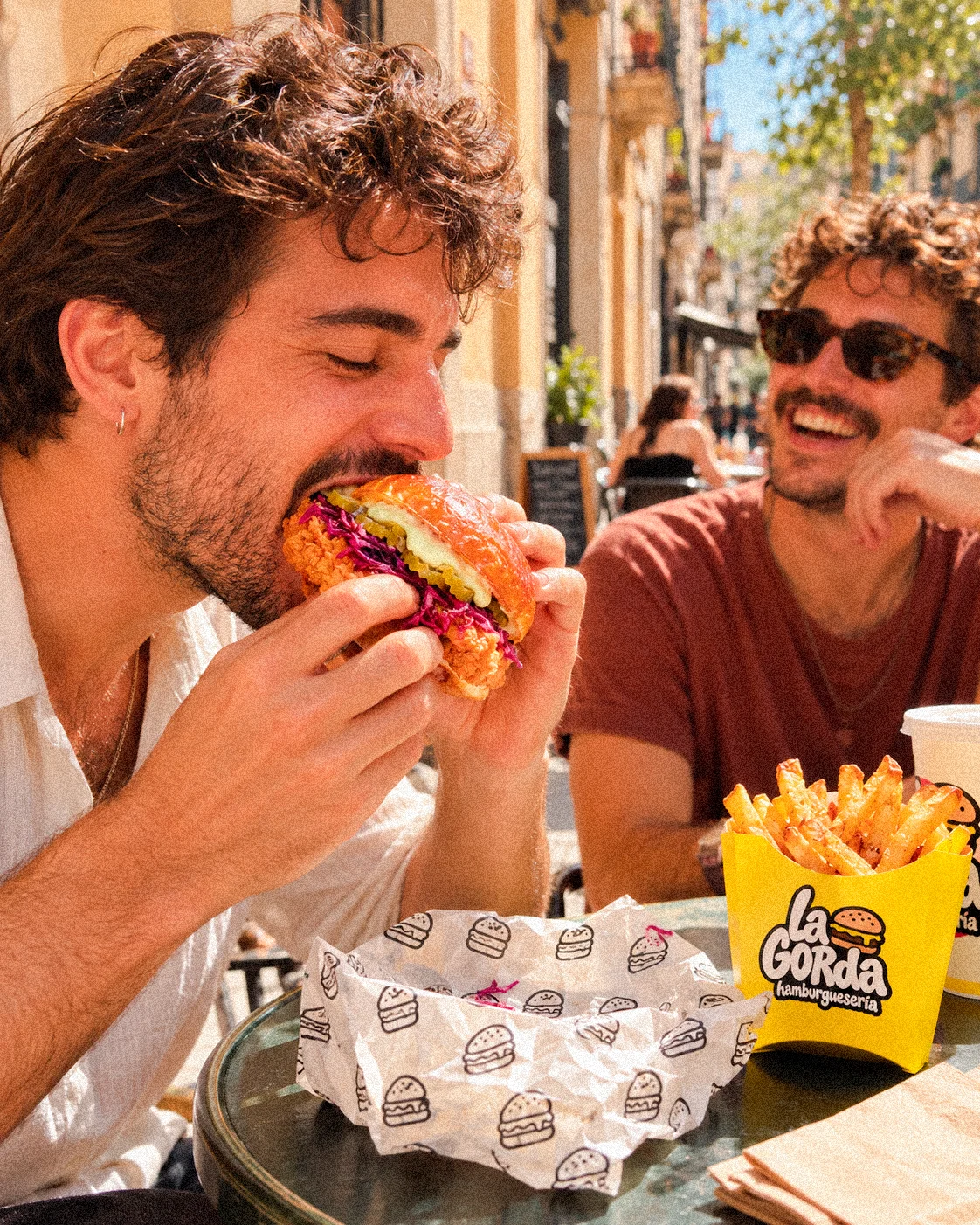 Dos amigos en una mesa de terraza; uno muerde una Pollo Turquesa, con patatas de La Gorda en la mesa