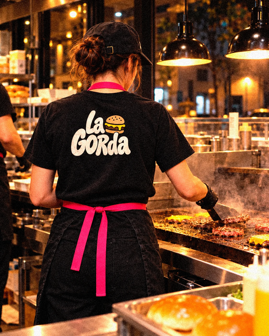 De noche, una cocinera de espaldas en la plancha con la camiseta negra de La Gorda y el mandil rosa